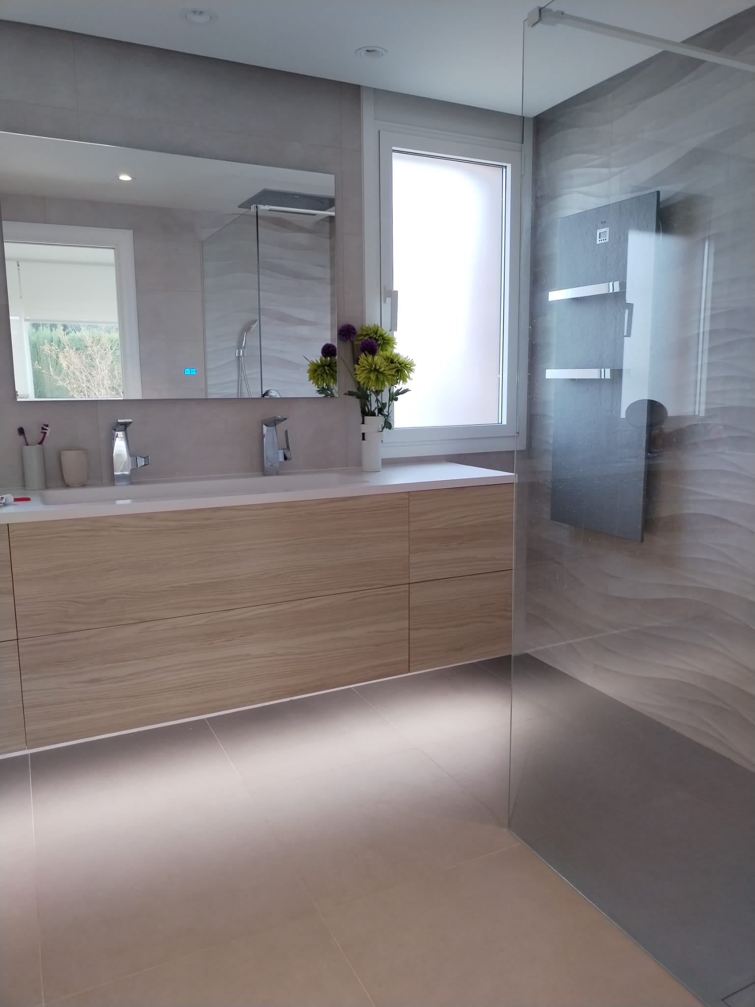 Baño moderno con lavabo doble, mueble de madera clara y ducha con pared de azulejos texturizados.