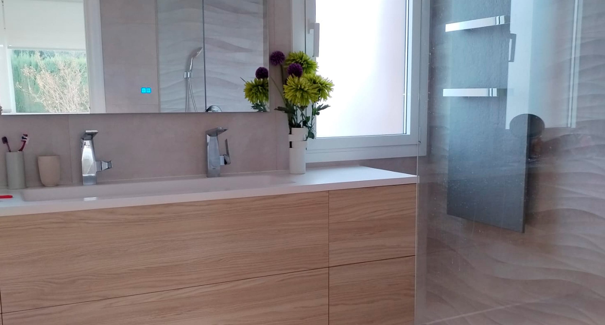 Detalle de baño moderno con lavabo doble, mueble de madera clara y ducha con pared de azulejos texturizados.