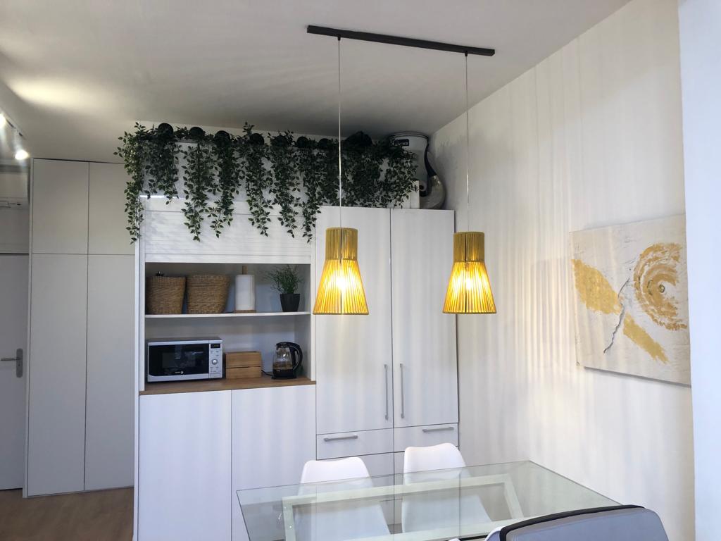 Comedor moderno con mesa de cristal, sillas blancas, lámparas colgantes amarillas y gabinetes blancos, con plantas colgantes decorativas en la parte superior y una pintura abstracta en la pared.